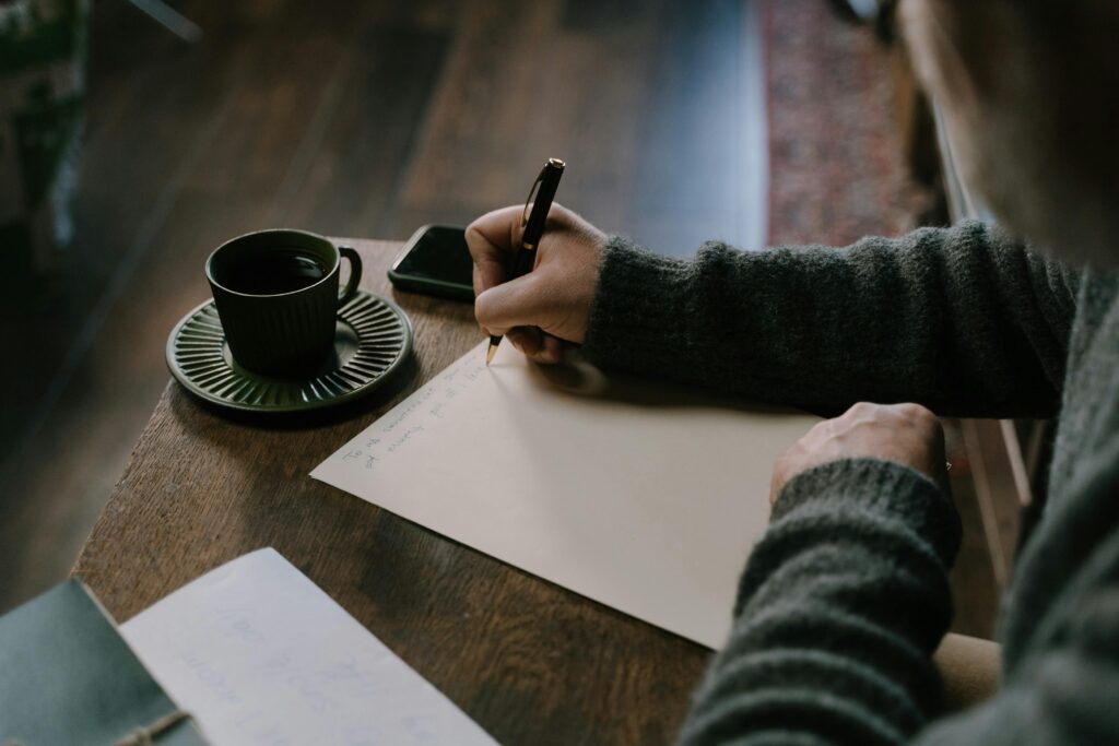 Close-up of a person writing on paper with a cup of coffee on the table.