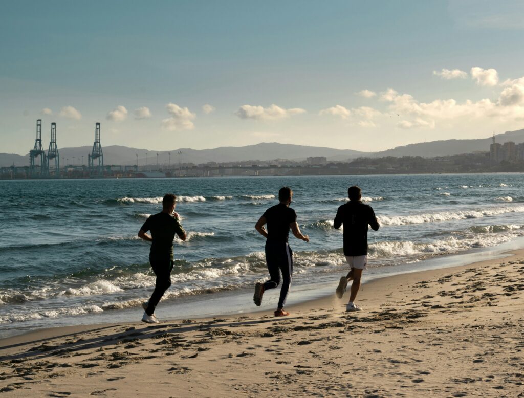 Three men jogging along a scenic beach, embracing a healthy lifestyle under a clear sky.