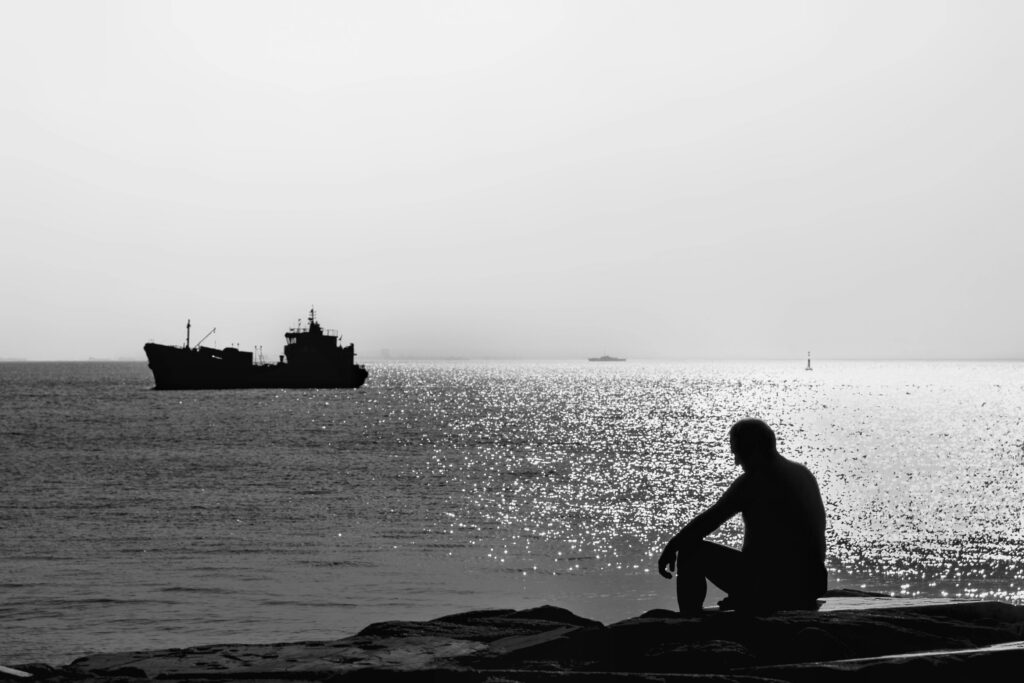Black and white silhouette of a man sitting by the sea with a ship in the distance.