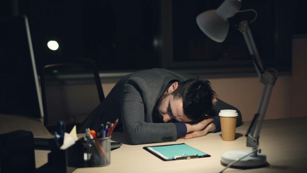 Man in office suit sleeping on desk with coffee and tablet nearby, symbolizing work fatigue.