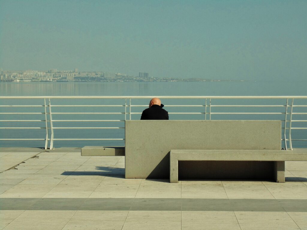 A solitary figure sitting on a bench overlooking a calm waterfront under a clear sky.